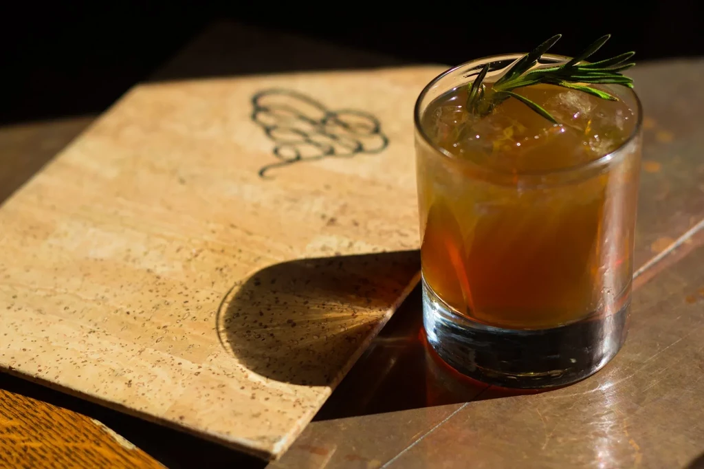 Reddish-brown cocktail with ice, rosemary, on dark table near cork.