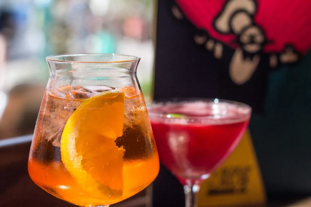 Amber-orange cocktail with orange slice, blurred red drink in background.