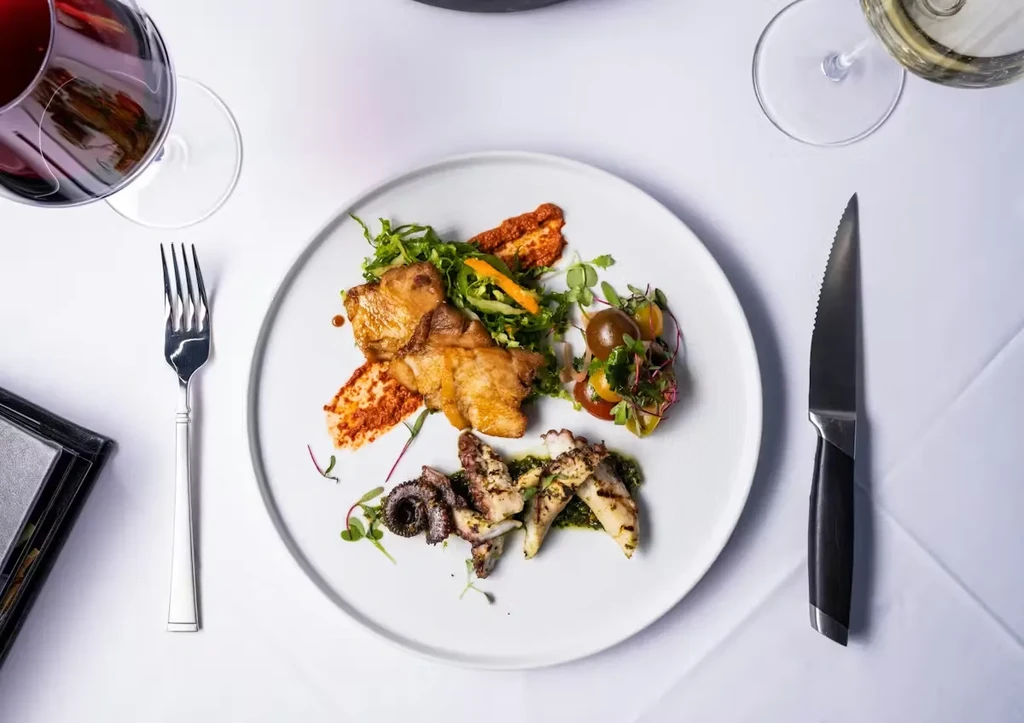 Elegant gourmet dish featuring meat, octopus, salad, and wine glasses.