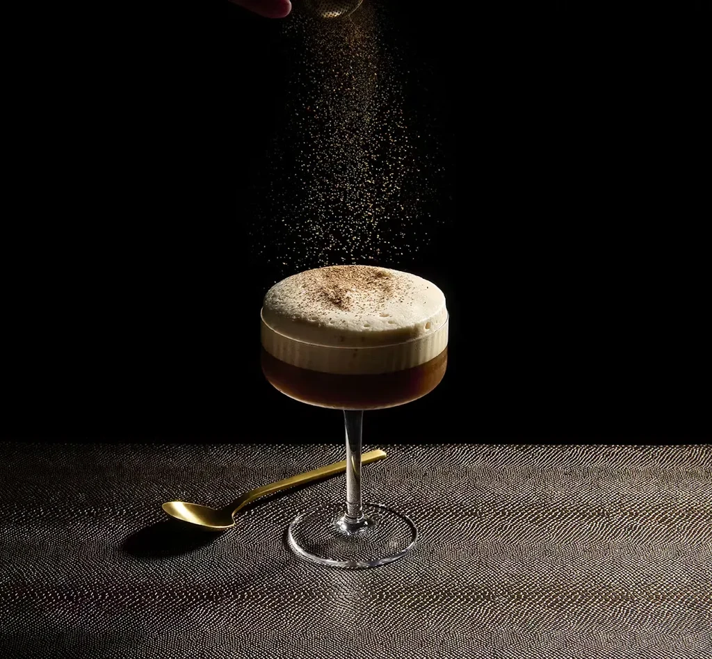 Layered drink in a stemmed glass, powder being sprinkled, golden spoon.