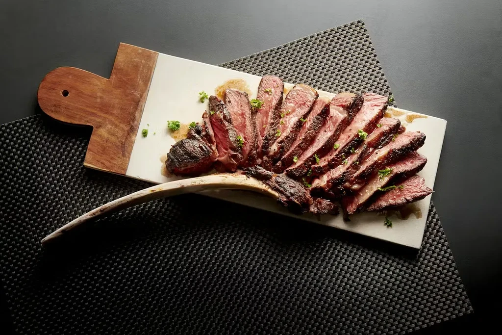 Sliced medium-rare tomahawk steak with herbs on a marble and wood board.