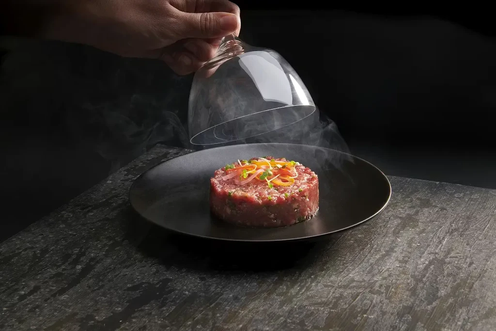 Hand lifts cloche, revealing smoking beef tartare on dark plate.