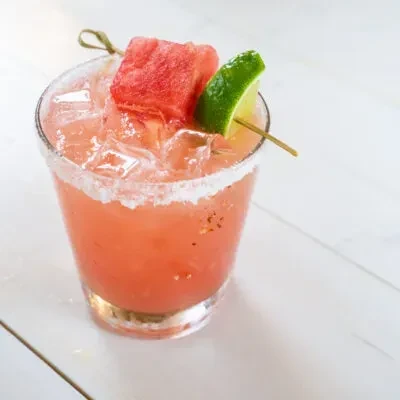 Coral drink with ice, salted rim, watermelon, and lime garnish.