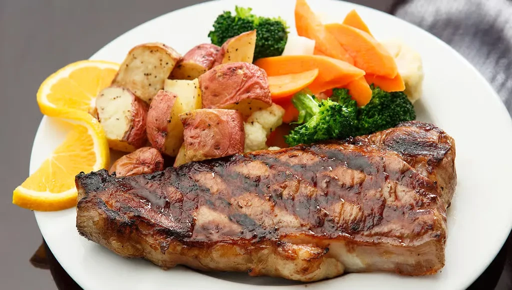 Grilled steak, roasted vegetables, and orange wedges on a white plate.