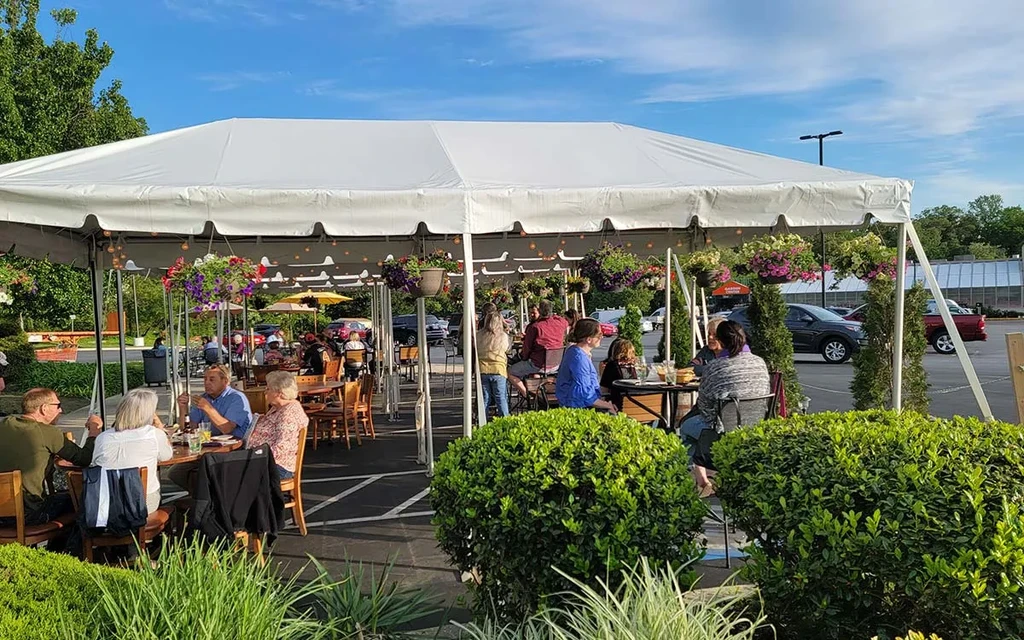 Lively outdoor restaurant patio under white tent with flowers and lights.