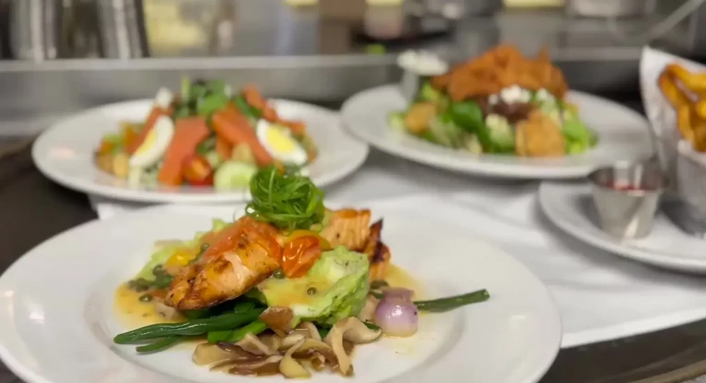Plates of grilled salmon, two salads, and fries on a kitchen pass.