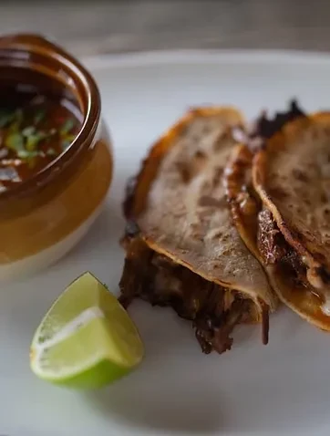 Three birria tacos with consommé, fresh herbs, and a lime wedge.