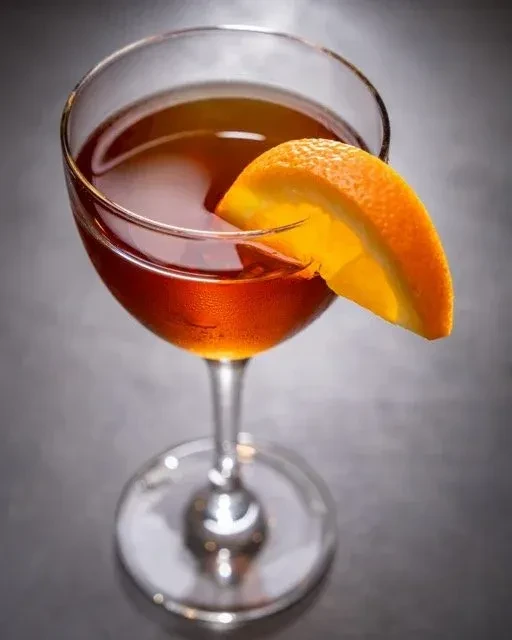 Amber cocktail in coupe glass with orange garnish, dark background.