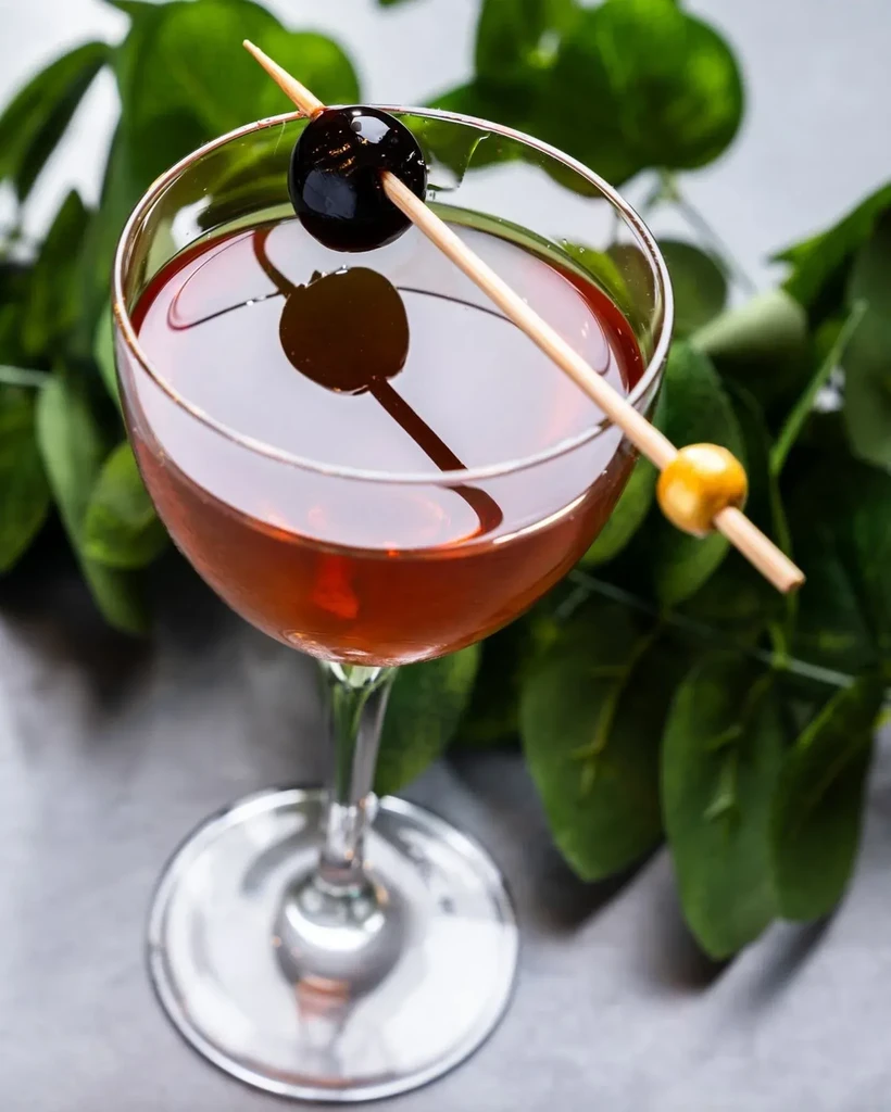 Amber cocktail in coupe glass with cherry garnish and green background.