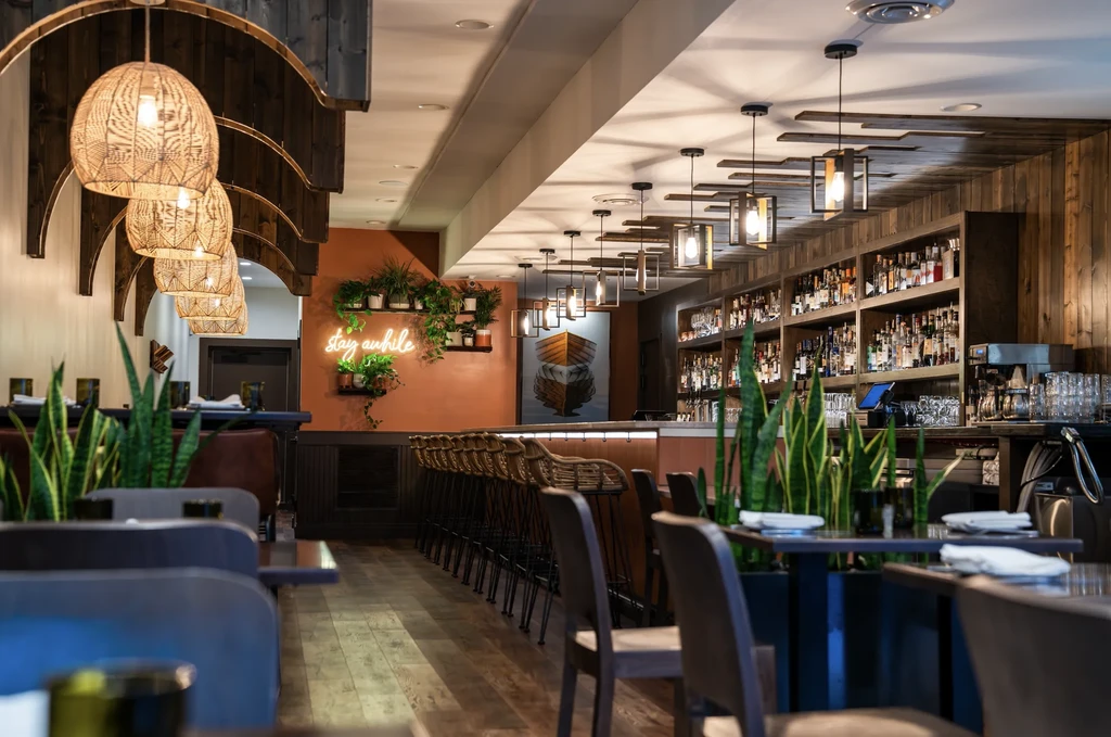 Modern restaurant and bar interior with natural wood, plants, and neon sign.