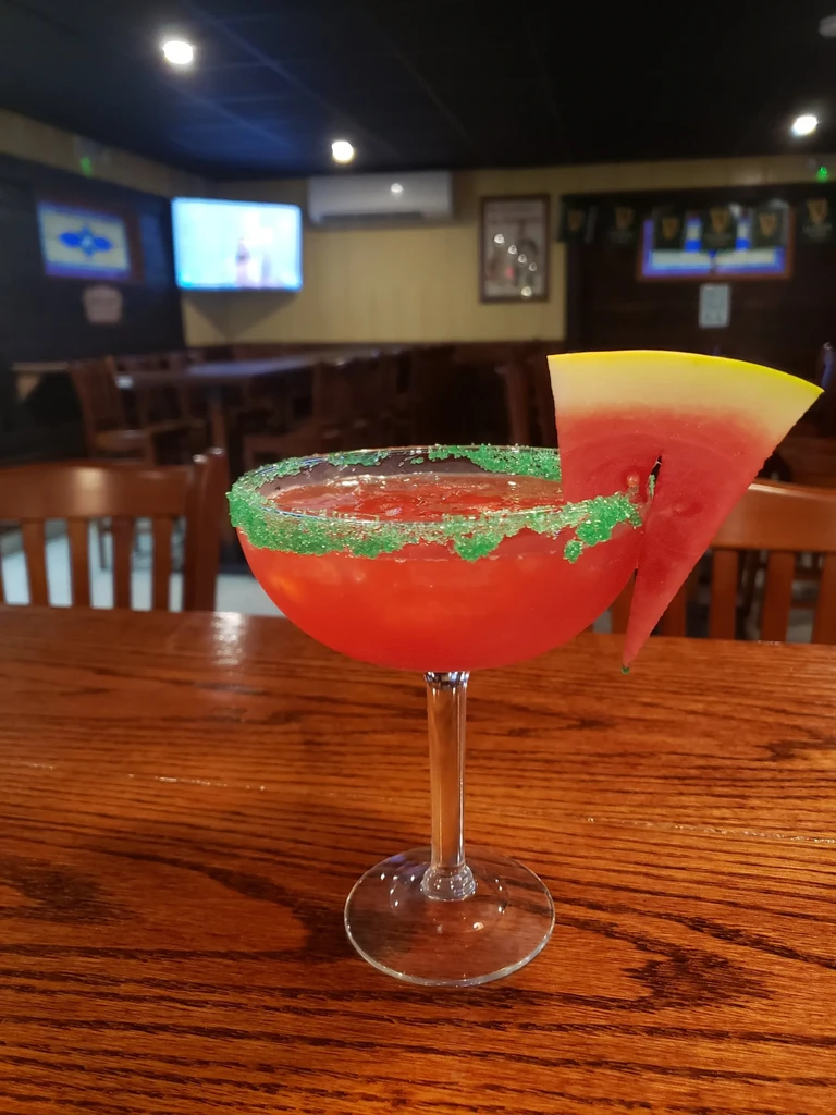 Red cocktail in margarita glass, green sparkly rim, watermelon garnish.