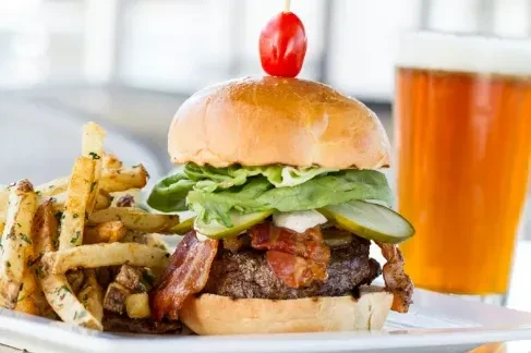 Burger with bacon, fries, and a beer on a white plate.