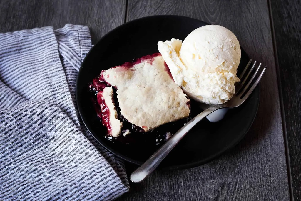 Berry pie slice with vanilla ice cream on black plate, wooden table.
