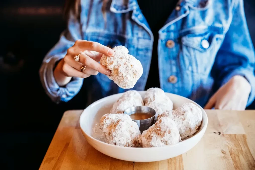 Hands picking a powdered sugar fritter from a bowl, with sauce.