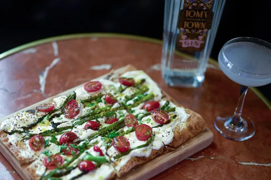 Rectangular flatbread with cheese, asparagus, tomatoes, gin bottle, and cocktail.