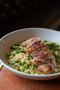 Seared duck breast over risotto with peas and asparagus in white bowl.
