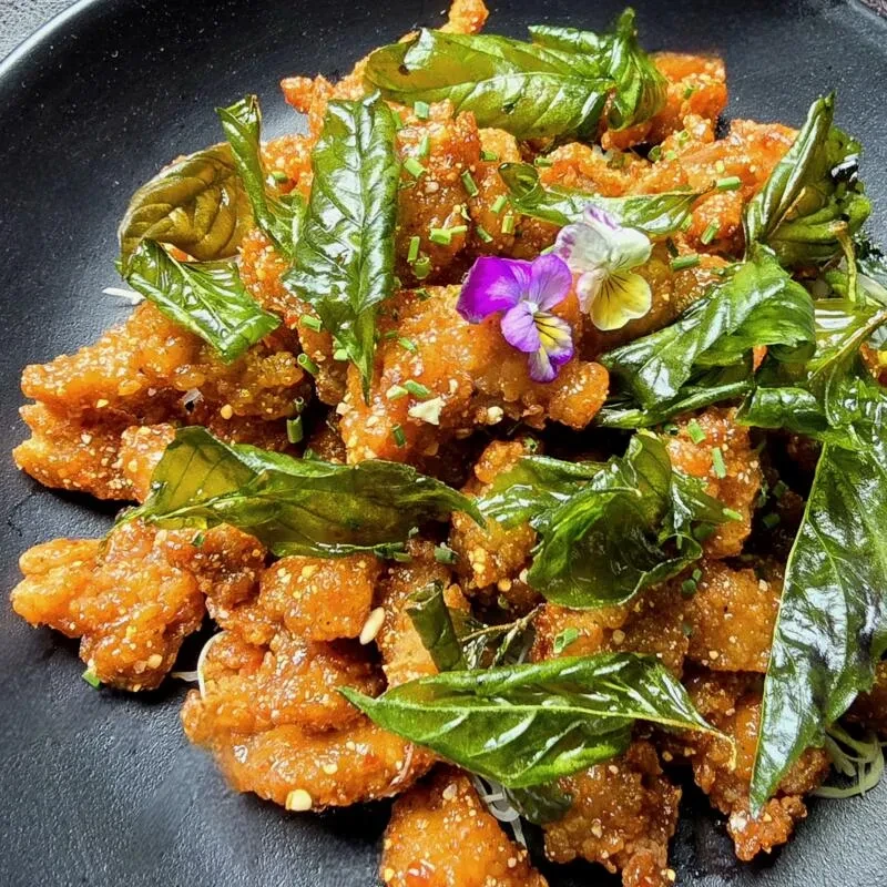 Crispy golden chicken with green herbs and edible flowers on black plate.
