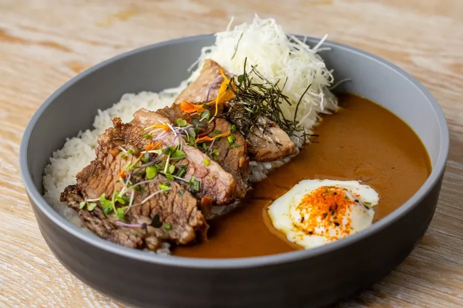 Garnished beef curry, rice, and soft-boiled egg in a dark bowl.