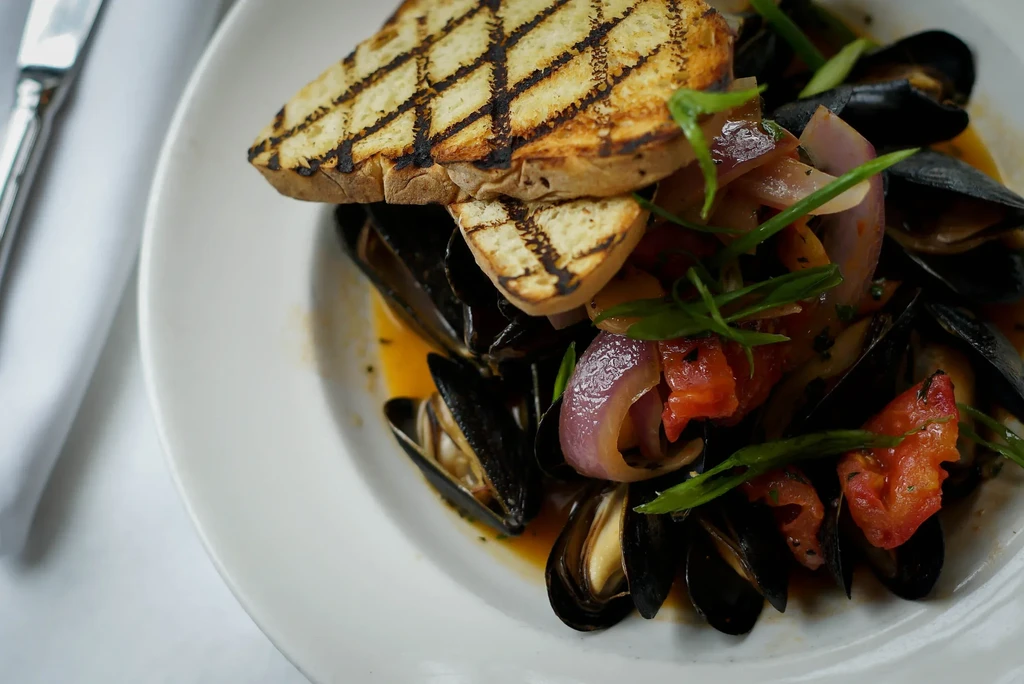 Mussels in reddish broth with grilled bread, onion, tomato, and herbs.