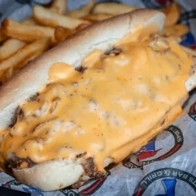 Cheesesteak sandwich, melted orange cheese, french fries on a branded liner.