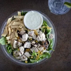 Plastic bowl of chicken salad with corn, tortilla strips, dressing, water.