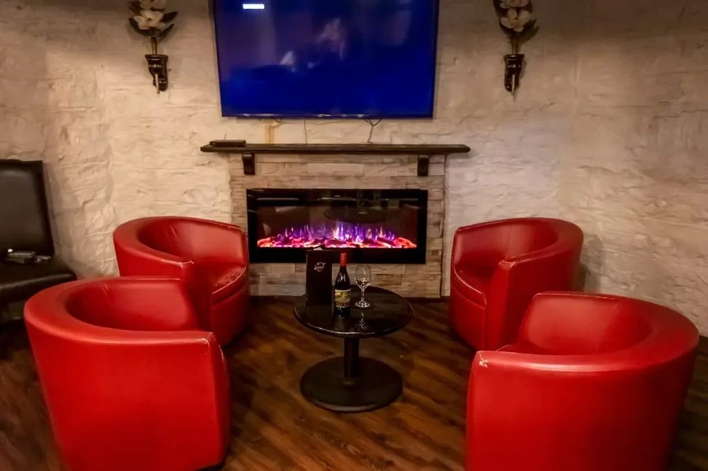 Lounge with electric fireplace, TV, red chairs, and wine.