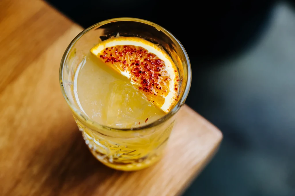 Light amber cocktail with ice and a chili-spiced orange slice.
