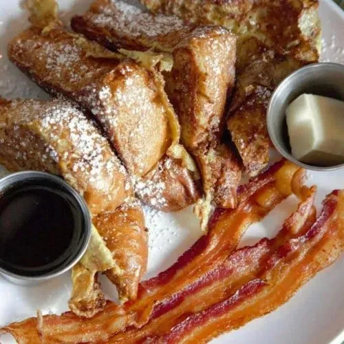 French toast sticks with powdered sugar, crispy bacon, syrup, and butter.
