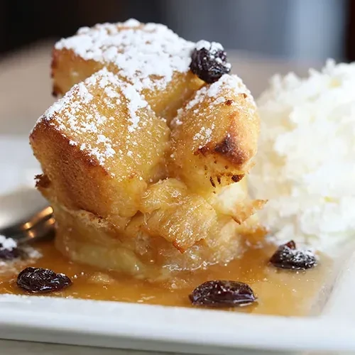 Golden-brown bread pudding with powdered sugar, raisins, amber sauce, and whipped cream.