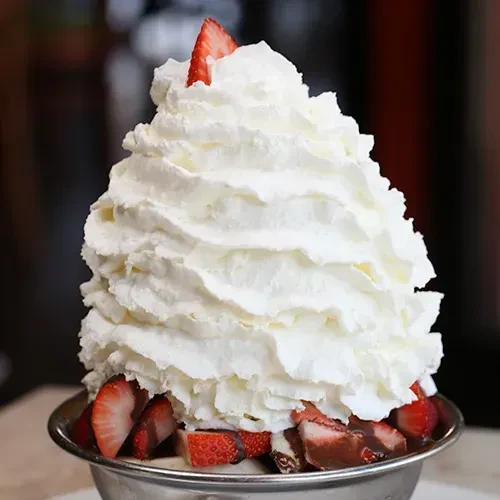 Dessert in silver bowl with whipped cream, strawberries, and dark sauce.