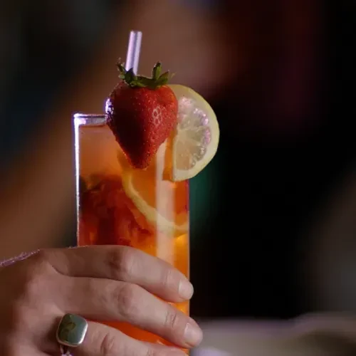 Hand with ring holding orange drink, garnished with strawberry and lemon.