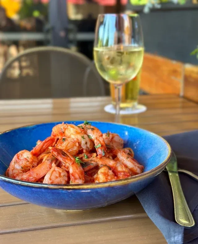 Vibrant blue bowl of shrimp with sauce, wine, and knife.