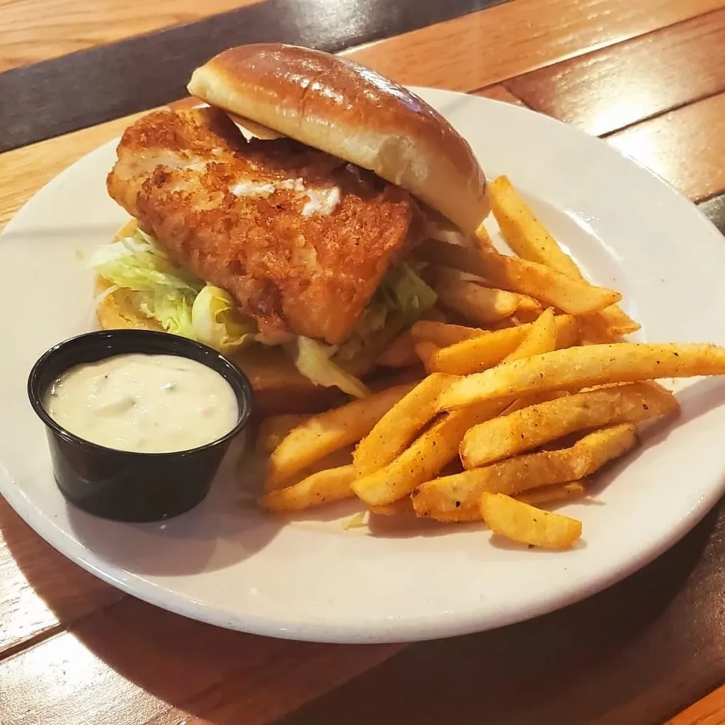 Crispy fried fish sandwich, french fries, and dipping sauce on a wooden table.