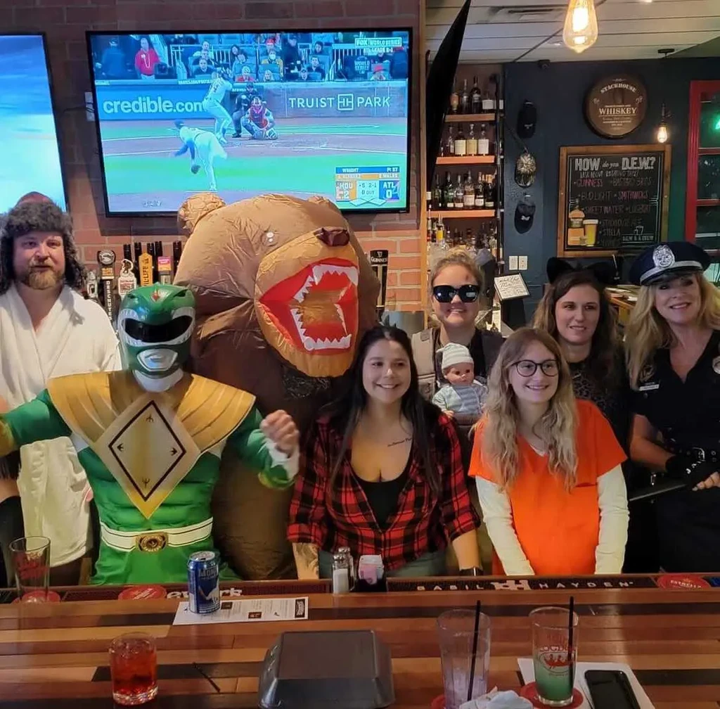 Eight costumed people pose in a bar with drinks and a TV.