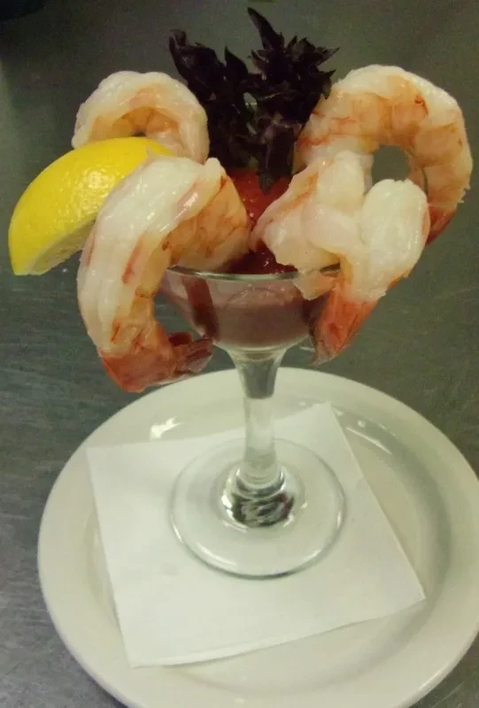Shrimp cocktail in a stemmed glass with red sauce, lemon, and herb.