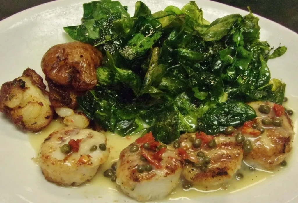 A close-up of seared scallops, roasted potatoes, and green leafy vegetables.