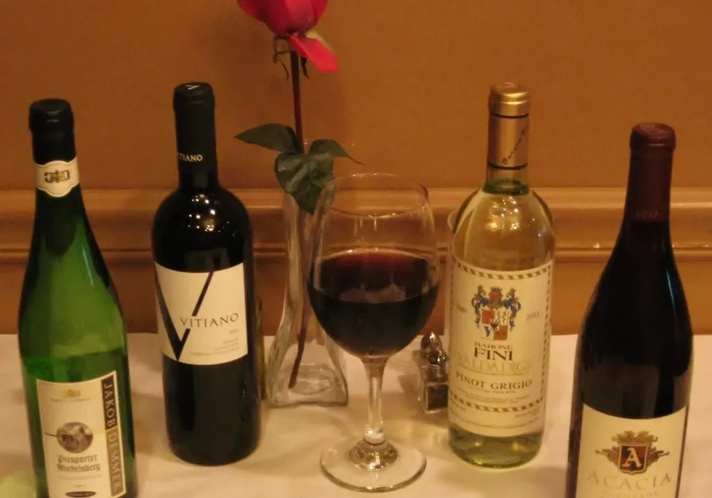 Four wine bottles, red wine glass, and red rose on table.