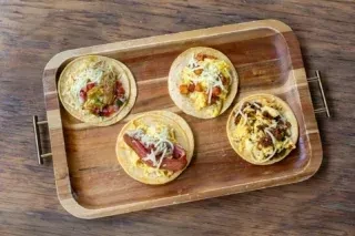 Four small, open-faced tacos with varied toppings on a wooden tray.
