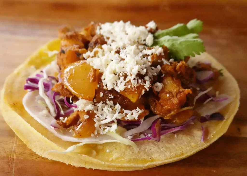 Single taco with meat, potatoes, cabbage, cheese, and cilantro on wood.