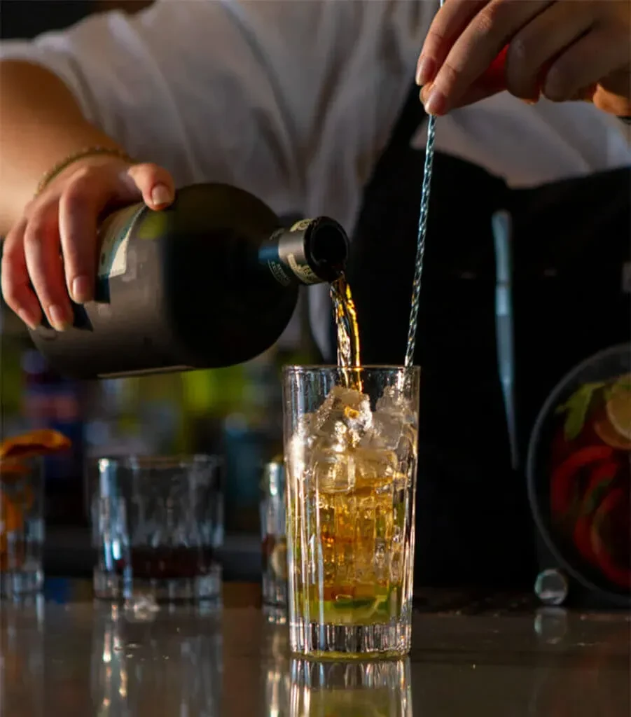 Bartender pours amber liquid into an iced glass, stirring.