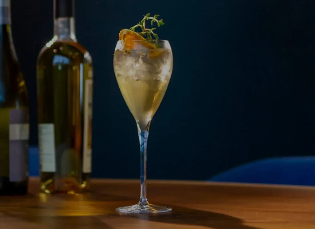 Amber cocktail in stemmed glass with citrus, herbs, on wood.