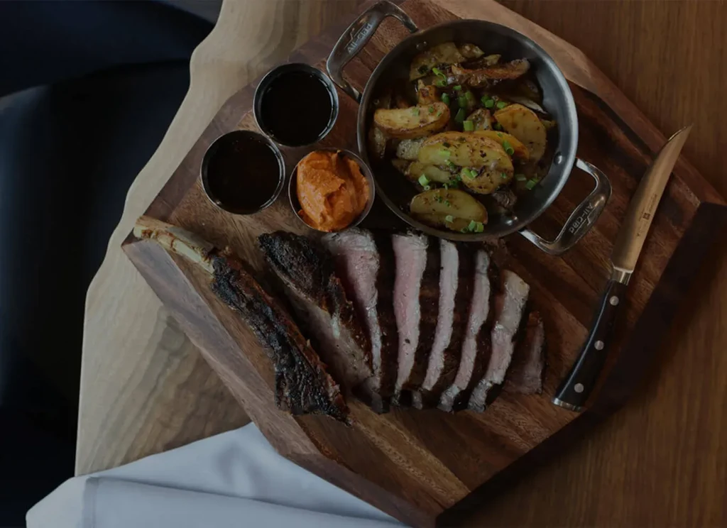 Sliced medium-rare steak, roasted potatoes, and sauces on wooden board.