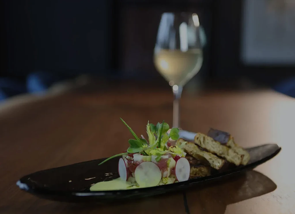 Plated gourmet dish with greens, toast, and white wine, dark setting.