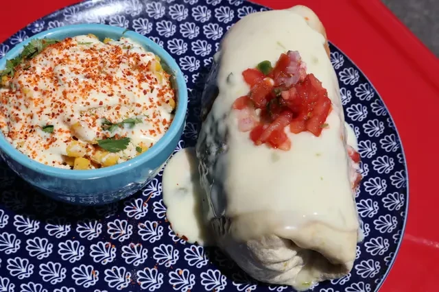 Burrito smothered in cheese sauce, with salsa and a side of esquites.