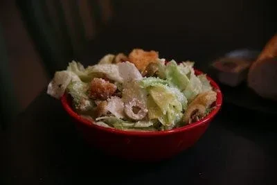 Dim-lit Caesar salad with lettuce, Parmesan, and croutons in a red bowl.