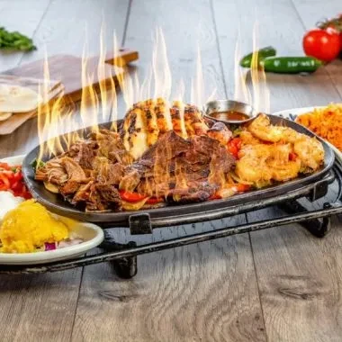 Sizzling cast iron platter of meats and vegetables with smoke and flames.