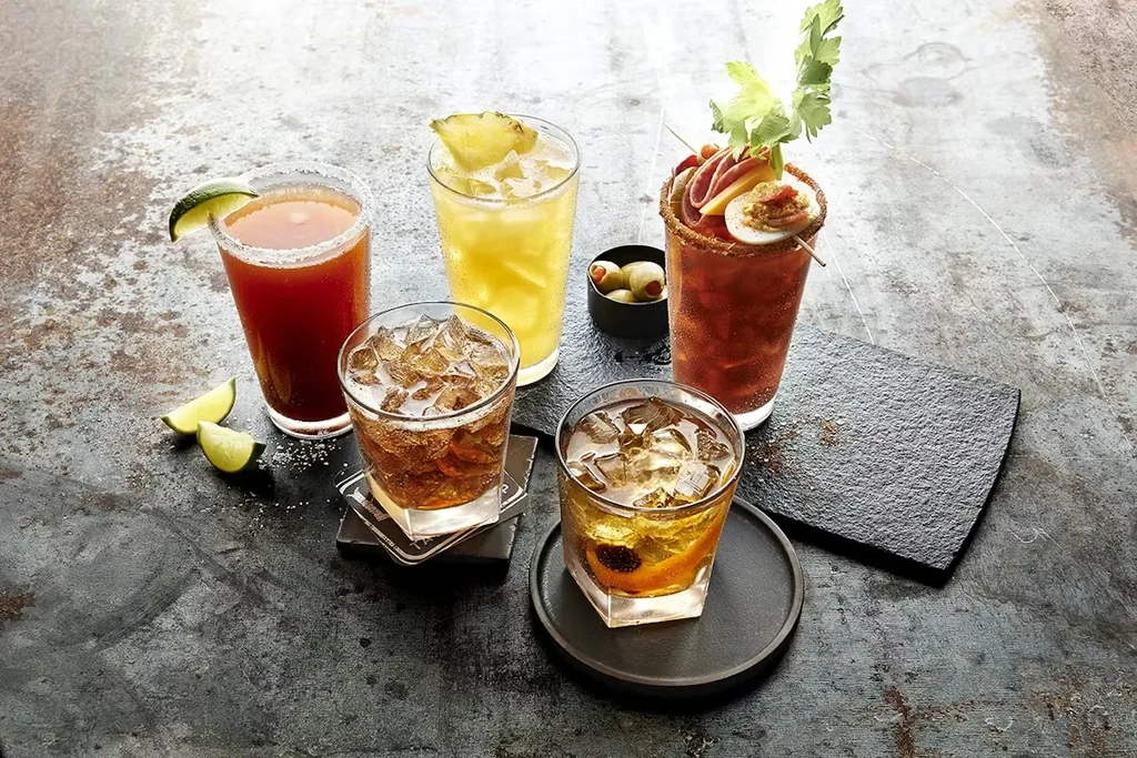 Five diverse cocktails, including a heavily garnished Bloody Mary, on a dark table.