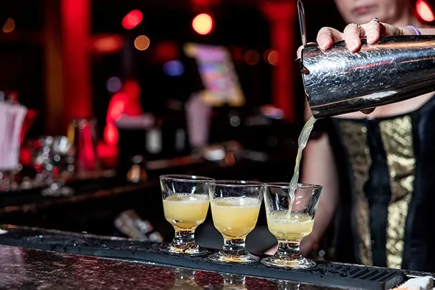 Bartender pours frothy yellow drink from shaker into glasses.