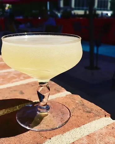 Pale yellow cocktail in a coupe glass on a sunny brick patio.