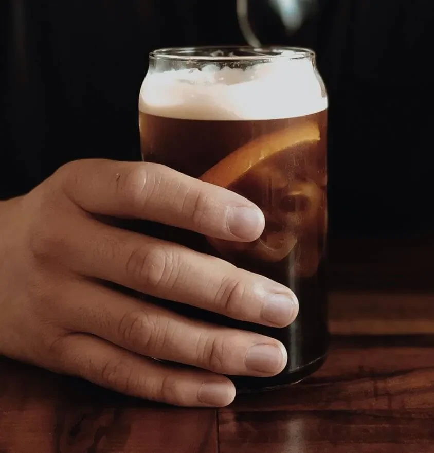 Hand holds dark foamy drink with orange in a can-shaped glass.